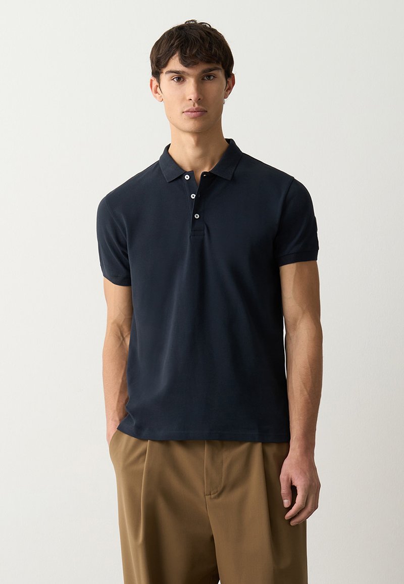 Polo shirt blu navy con una texture morbida, maniche corte, colletto classico e patta con tre bottoni. Abbinato a pantaloni beige larghi.