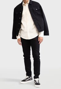 Svart denimjacka över en beige skjorta med knappar, kombinerad med svarta slim-fit byxor och svarta sneakers med vita accenter.