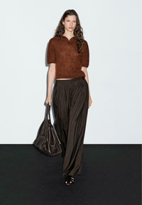 Bruine gebreide polo trui met korte mouwen, zwarte geplooide broek en een grote zwarte leren tas, tegen een lichte achtergrond.
