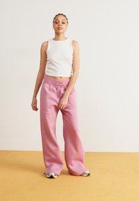 Witte geribbelde tanktop gecombineerd met roze sweatpants met een trekkoord in de taille, een ontspannen pasvorm en een gladde textuur. Geplaatst tegen een neutrale achtergrond.