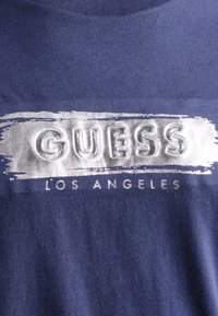 Camiseta de algodón azul marino con un logo impreso en plata que dice "GUESS LOS ÁNGELES" sobre un fondo texturizado de pinceladas.