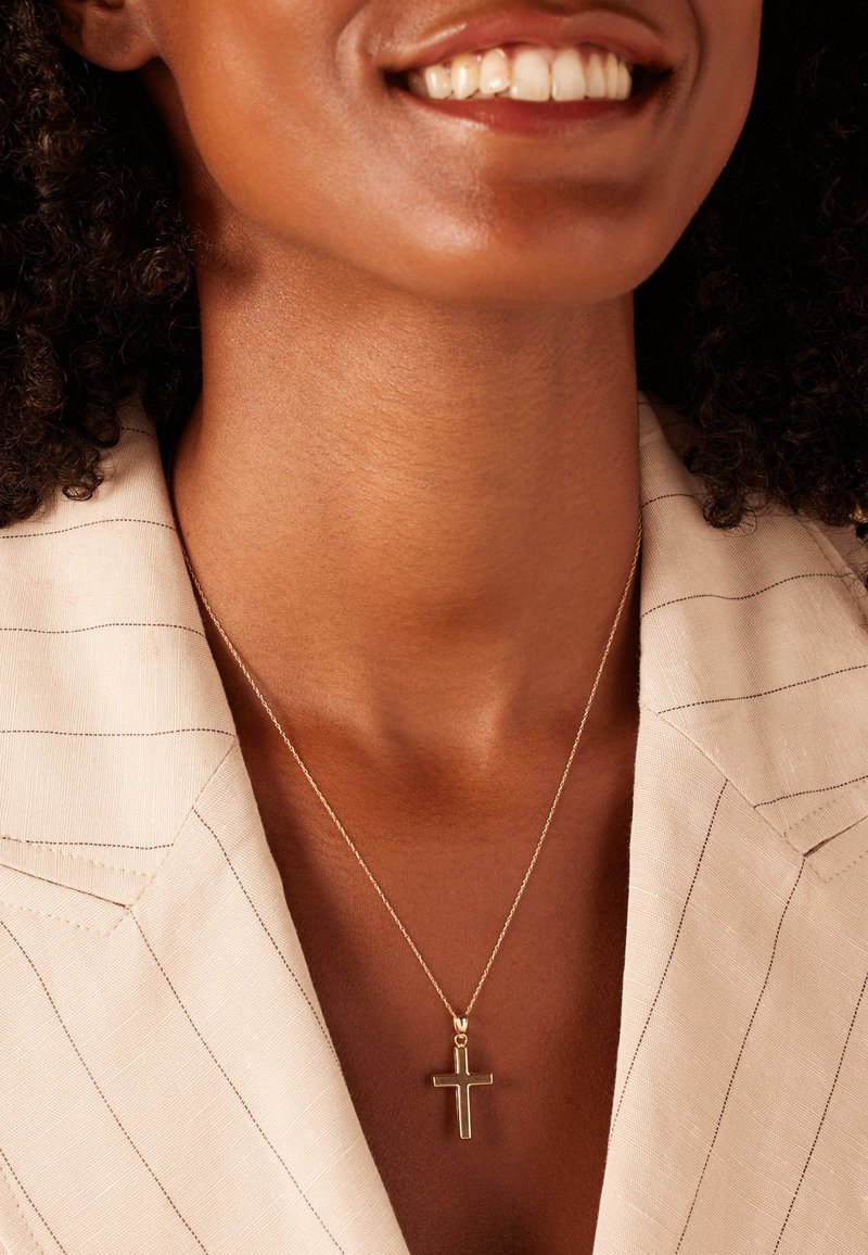 Gouden kruis ketting aan een dunne ketting, gedragen over een lichtgekleurde, gestreepte blouse, die een gladde hals en een eenvoudig ontwerp benadrukt.