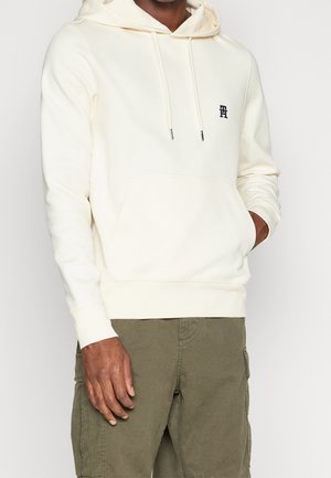 Sweatshirt à capuche crème avec poche kangourou sur le devant, ourlet et poignets côtelés, et un petit logo brodé sur la poitrine.