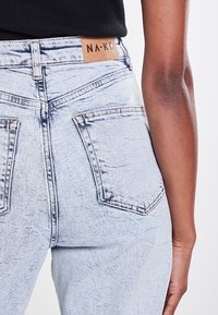 Jean en denim bleu clair avec un délavage, deux poches arrière et une étiquette en cuir beige avec "NA-KD" cousue à la taille.