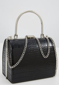 Musta krokotiilikuvioitu nahkainen käsilaukku hopeisella metallikehyksellä, kaarevalla kahvalla ja hopeaketjuhihnalla valkoisella pinnalla.
