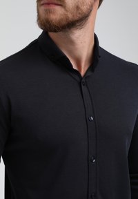 Camisa negra de botonadura con una textura sutil, presenta un collar de punta y botones oscuros en la parte delantera. Diseño ajustado.