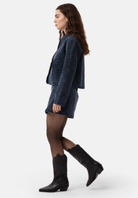 Chaqueta texturizada en azul marino y falda a juego, con botas de vaquero negras hasta la rodilla. La modelo está de lado contra un fondo blanco.