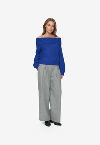 Pull bleu à épaules dénudées en tricot texturé, associé à un pantalon large taille haute gris. Des chaussures noires complètent le look.