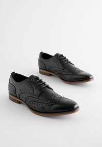 Must mustad brogue'de nahkasaapad keerukate perforatsioonide, terava nina ja madala puidust kontsaga. Tekstureeritud pealmine osa kontrastsete aktsentidega.