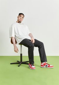 Ung man med vitiligo iklädd en vit t-shirt, svarta jeans och röd-vit sneakers sitter på en snurrstol på ett grönt golv mot en vit vägg.