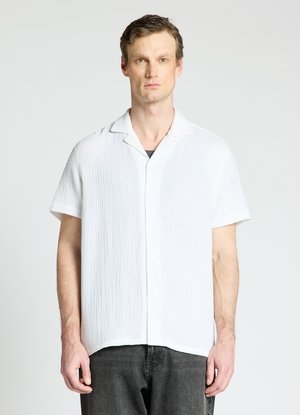 Homme portant une chemise blanche texturée à manches courtes boutonnée, avec un pantalon noir, debout devant un fond clair uni.
