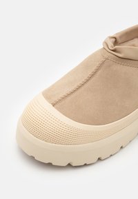 Beige Wildledersneaker mit einer dicken, cremefarbenen Gummisohle, strukturierten Akzenten und einem lässigen elastischen Kragen für einen sicheren Sitz.
