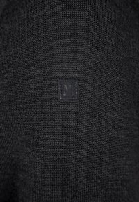 Schwarzer Strickpullover mit einem kleinen quadratischen Etikett mit dem Buchstaben "M". Die Stoffoberfläche ist durchgehend glatt und gleichmäßig.