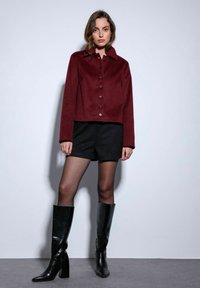 Bordeauxrode cropped jas met een kraag en zwarte knopen. Gecombineerd met zwarte shorts en kniehoge, puntige zwarte laarzen. Gladde textuur, modern ontwerp.