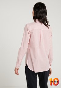 Vrouw met donker golvend haar draagt een langemouwen lichtroze shirt met fijne rode strepen, gecombineerd met zwarte broek, staande tegen een effen achtergrond.
