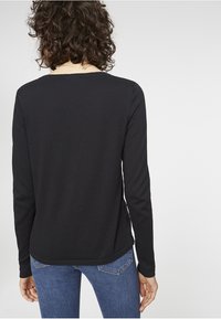 Person med lockigt hår som bär en långärmad svart tröja och blå jeans, stående vänd bort från kameran mot en enkel ljus bakgrund.