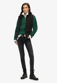 Svart ärmlös väst kombinera med en grön kabelstickad tröja, vit skjorta med krage, svarta smala jeans och svarta snörade kängor.