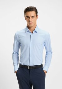 Camicia a maniche lunghe blu chiaro con colletto a bottoni e righe verticali, abbinata a pantaloni scuri e una cintura nera. Tessuto morbido.