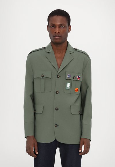Veste militaire verte au style décontracté, dotée de plusieurs poches, de boutons et de divers écussons à l'avant. Tissu texturé.