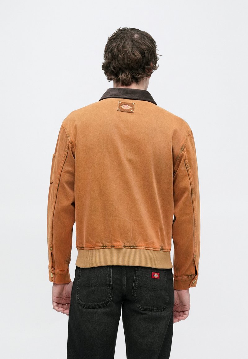 Brown corduroy jacket with a dark collar, ribbed hem, and buttoned pockets, featuring a stitched label at the back. Worn over dark jeans.