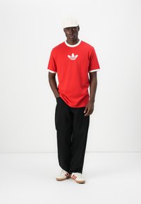 Κόκκινο κοντομάνικο μπλουζάκι με λευκές ραφές και κεντρικό λευκό γραφικό λογότυπο, σε συνδυασμό με μαύρο φαρδύ παντελόνι και λευκά αθλητικά παπούτσια.