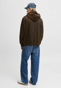 Brown hooded sweatshirt with a relaxed fit, paired with loose blue jeans. The person is wearing a light blue cap and white sneakers.