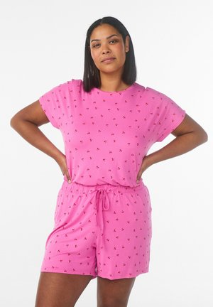 Femme à la peau moyen-foncée et aux cheveux noirs portant un haut à manches courtes rose à motifs de cerises et un short assorti, debout les mains sur les hanches.