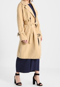 Trench-coat beige à manches longues, taille ceinturée et poches latérales. Associé à des culottes bleu marine et des sandales à talons noires.