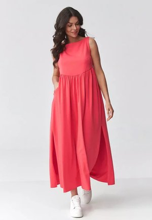 Kobieta w koralowej, bezrękawnikowej sukience maxi i białych trampkach, idąca z rękami w kieszeniach sukienki na jednolitym tle.