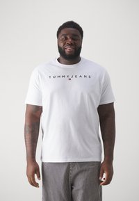 Balta medvilninė marškinėliai su juodu užrašu "TOMMY JEANS" per krūtinę; apvalus iškirpimas, trumpomis rankovėmis, suderinti su dryžuotais kelnėmis.