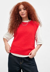 Junge Frau mit langen lockigen roten Haaren trägt einen leuchtend roten ärmellosen Pullover über einem weißen Hemd und dunklen Jeans und posiert mit den Händen am Saum.