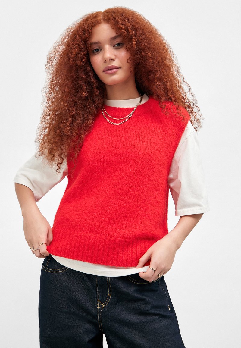Junge Frau mit langen lockigen roten Haaren trägt einen leuchtend roten ärmellosen Pullover über einem weißen Hemd und dunklen Jeans und posiert mit den Händen am Saum.