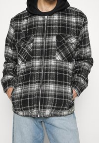 Hombre con chaqueta de cuadros en blanco y negro con cierre de cremallera, bolsillos con solapa en el pecho y manos en los bolsillos laterales, combinado con vaqueros azul claro.