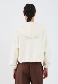 Korte crèmekleurige hoodie van zachte stof met een trekkoordcapuchon, elastische zoom en geribbelde mouwen, gedragen met bruine broeken. Minimalistisch ontwerp.