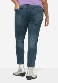 Vaqueros de mezclilla azul ajustados con bolsillos traseros y un ligero desgaste, combinados con botines plateados metálicos y una blusa lavanda.