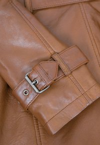 Brauner Lederjackenärmel mit einem Schnallen-Detail, das eine glatte Textur und sichtbare Nähte auf einem helleren braunen Hintergrund zeigt.