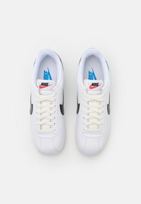 Sneaker Nike bianche con una finitura in pelle liscia, swoosh nero, lacci bianchi e un accento blu sulla soletta. Design classico a basso profilo.