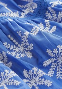 Tessuto blu con motivi floreali ricamati bianchi, con una finitura morbida e TEXTURATA e dettagli in pizzo decorativi nella parte superiore.