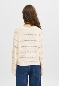 Krämsfärgad stickad tröja med hålmönster, ribbad fåll och långa ärmar. I kombination med blå denimjeans, sedd från baksidan.