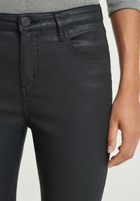 Svarta jeans i denim med en slät yta, med knappstängning, bälteslitsar och framfickor. Närbild av passform och design detaljer.