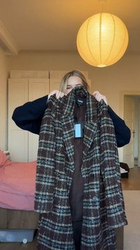 Brauner karierten langer Mantel mit blauen und weißen Akzenten, ausgestattet mit einem Reverskragen und Knöpfen. Präsentiert in einem hellen Innenraum.