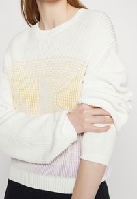 Personne portant un pull en tricot blanc avec des bandes horizontales jaunes et violettes, remontant la manche d'un bras.