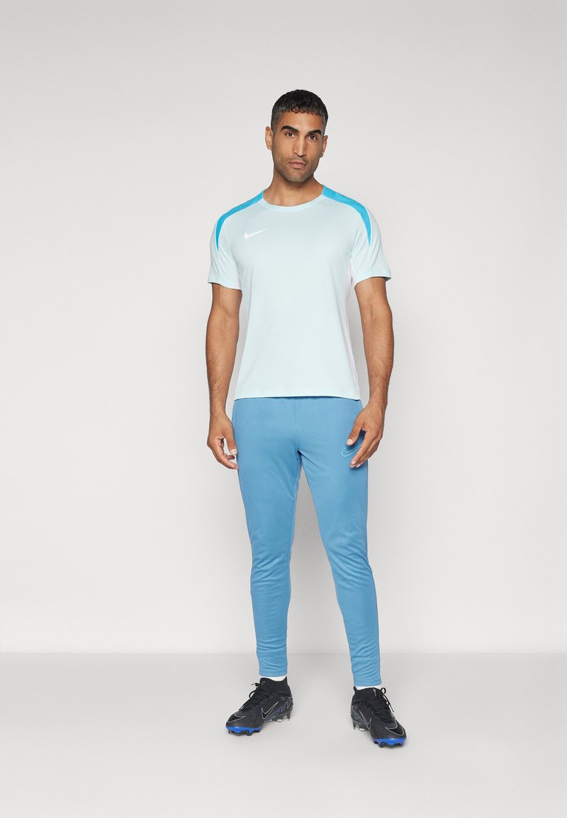 Mann in hellblauem und weißem Sporthemd, blauen Sporthosen und schwarzen Sportschuhen steht vor einem schlichten weißen Hintergrund.