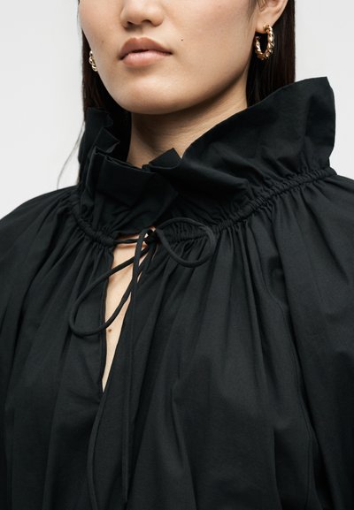 Γυναίκα που φοράει μαύρο πουκάμισο με βολάν στον ψηλό γιακά και δεμένη λαιμόκοψη, με αξεσουάρ χρυσά στριφτά κρίκους σκουλαρίκια.