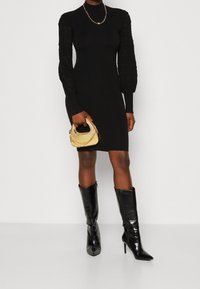 Sort tætsiddende kjole med teksturerede, puffede lange ærmer. Kombineret med skinnende sorte knæhøje støvler og en lille guldfarvet håndtaske med kæde-detaljer.