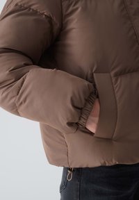 Brown padded jacket with elastic cuffs, featuring a side pocket and a drawstring detail at the hem, crafted from smooth, durable fabric.