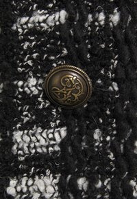 Stickad tyg i svart och vitt, med en framträdande, dekorativ bronsspänne med intrikata mönster. Texturen är fluffig och lagerpålagd.