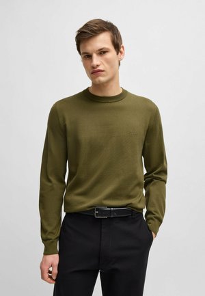 Olive green knitted sweater with long sleeves and a round neck, featuring a subtle logo detail on the chest, paired with black pants and a belt.