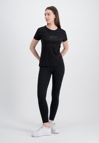 Schwarzes T-Shirt mit kurzen Ärmeln, das mit einem aufgedruckten Logo und Text versehen ist, kombiniert mit schwarzen Leggings und weißen Sneakern vor einem hellen Hintergrund.