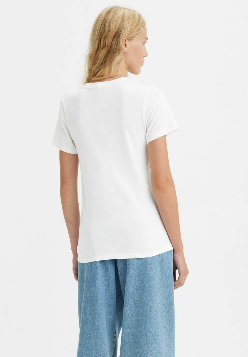 Femme aux longs cheveux blonds portant un t-shirt blanc à manches courtes unis et un jean léger bleu clair, de dos contre un fond blanc.